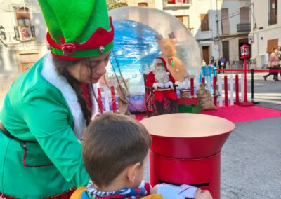 Niño entregando su carta a Papá Noel en un buzón rojo, asistido por un elfo.