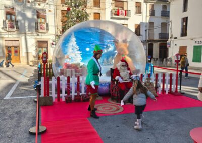 Niña saludando a Papá Noel junto a un duende en una bola de nieve gigante.