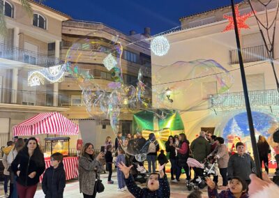 Niños jugando con burbujas gigantes en una plaza iluminada durante un evento festivo.