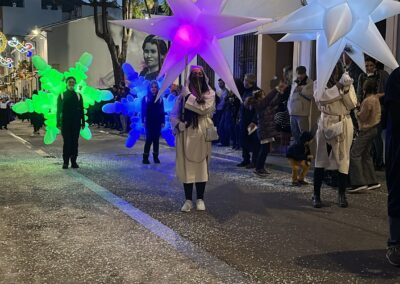 Pasacalles nocturno con elementos hinchables luminosos en una calle iluminada por decoraciones navideñas.