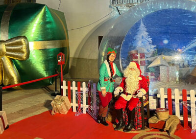 Santa Claus y su ayudante en una decoración navideña con un regalo inflable gigante y una esfera navideña.